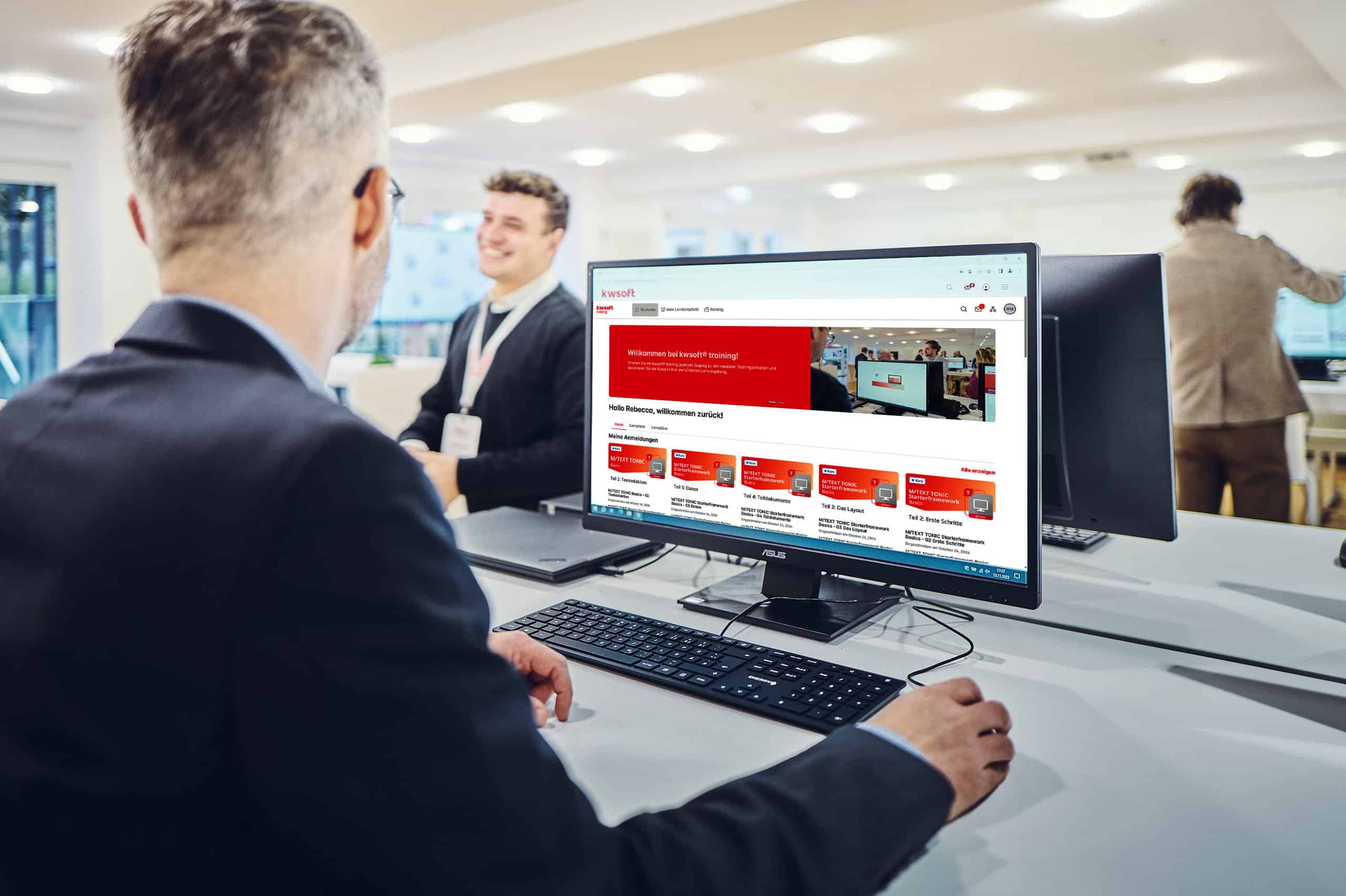 Un hombre está sentado frente a una pantalla en la que se muestra una formación de kwsoft y mira a un hombre que está de pie al fondo.
